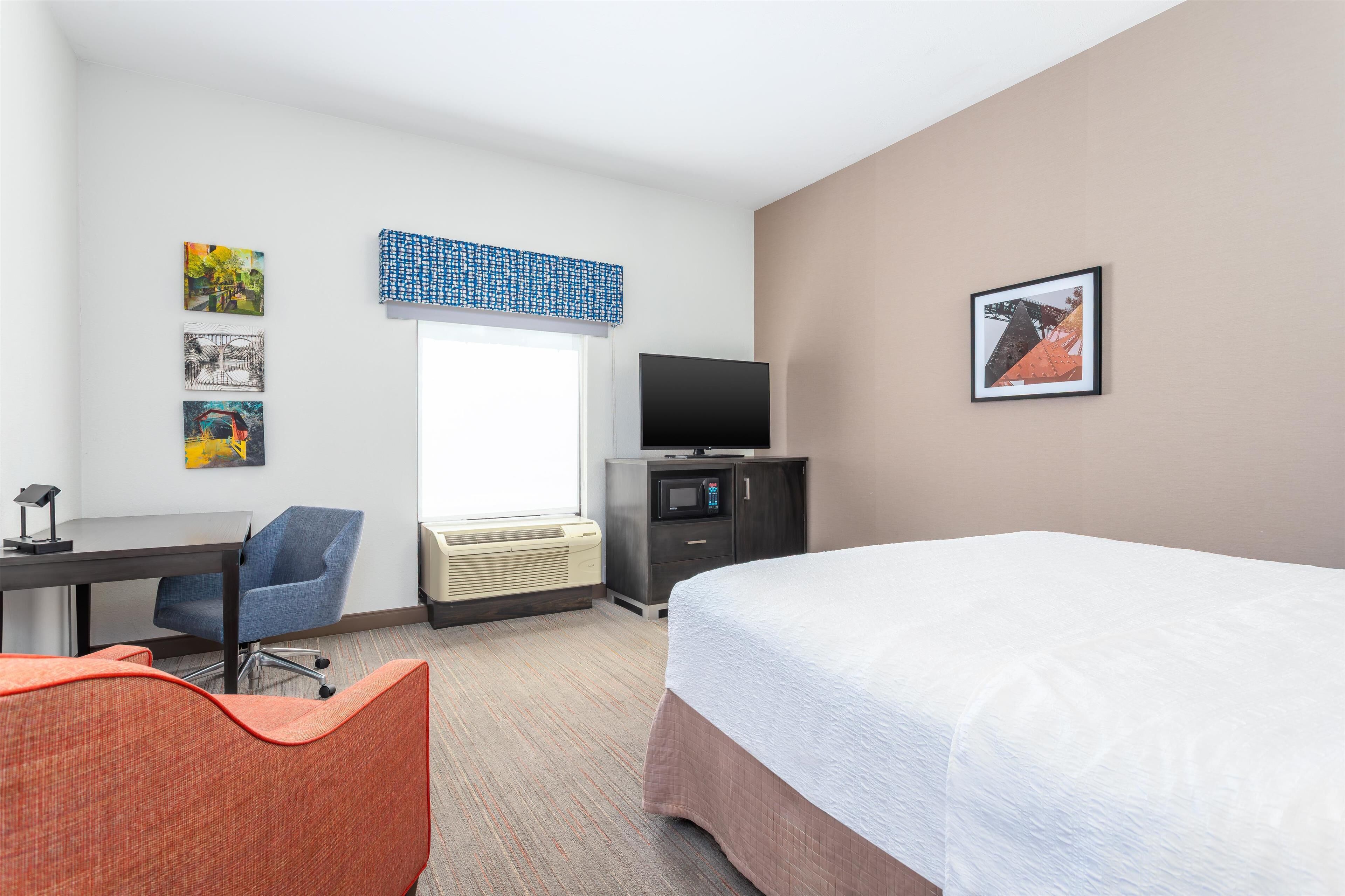 A hotel room with a bed, a desk with a chair, a television, and artwork on the walls.