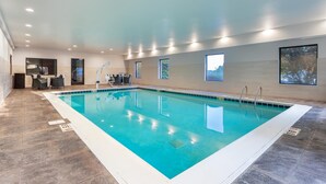 A large indoor swimming pool with clear blue water, surrounded by a tiled floor and walls. There are windows along the walls, and a seating area with tables and chairs is visible in the background. - Hampton Inn Akron-South (Akron)
