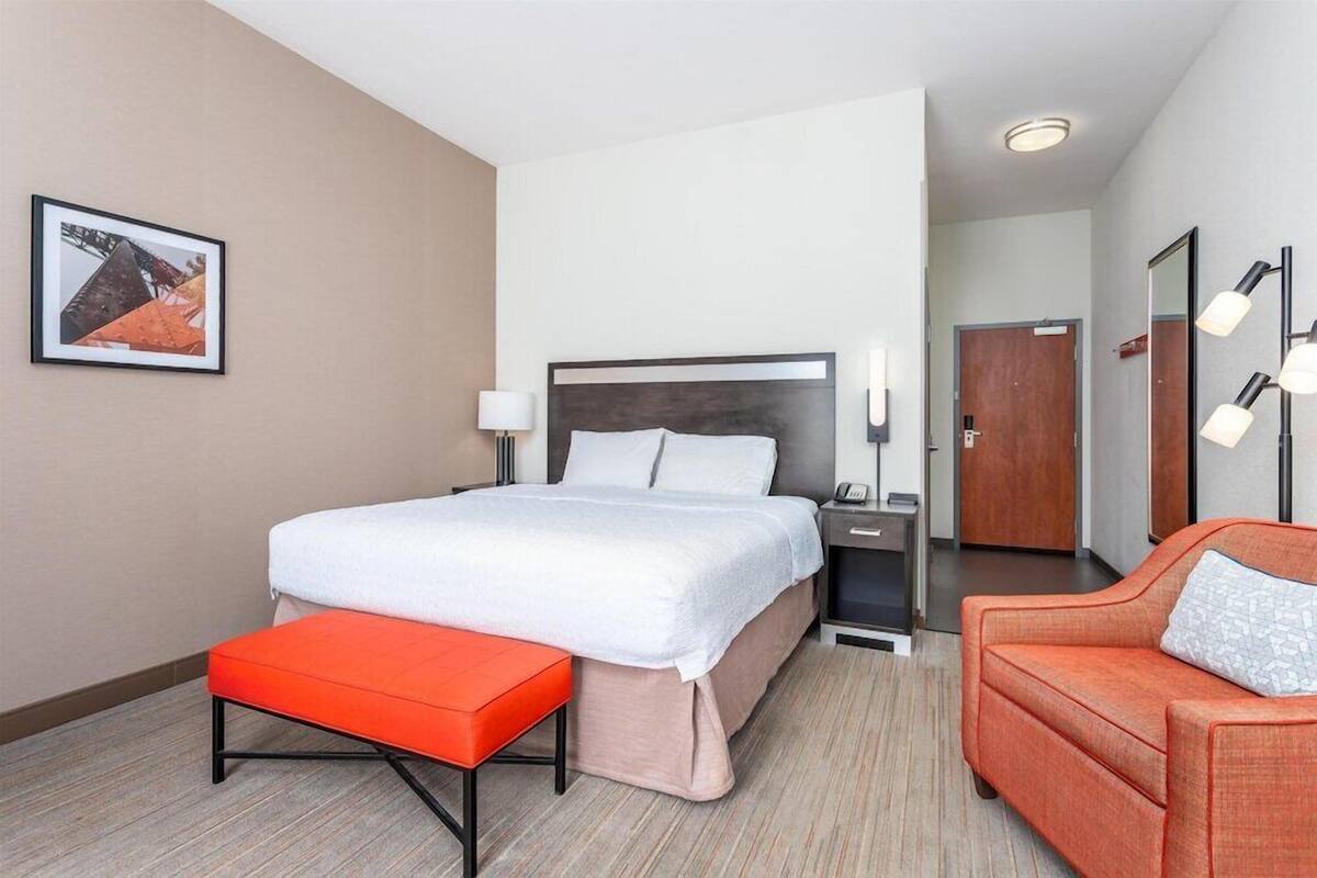 A hotel room with a bed, a nightstand, a red bench, a sofa, and a framed picture on the wall.