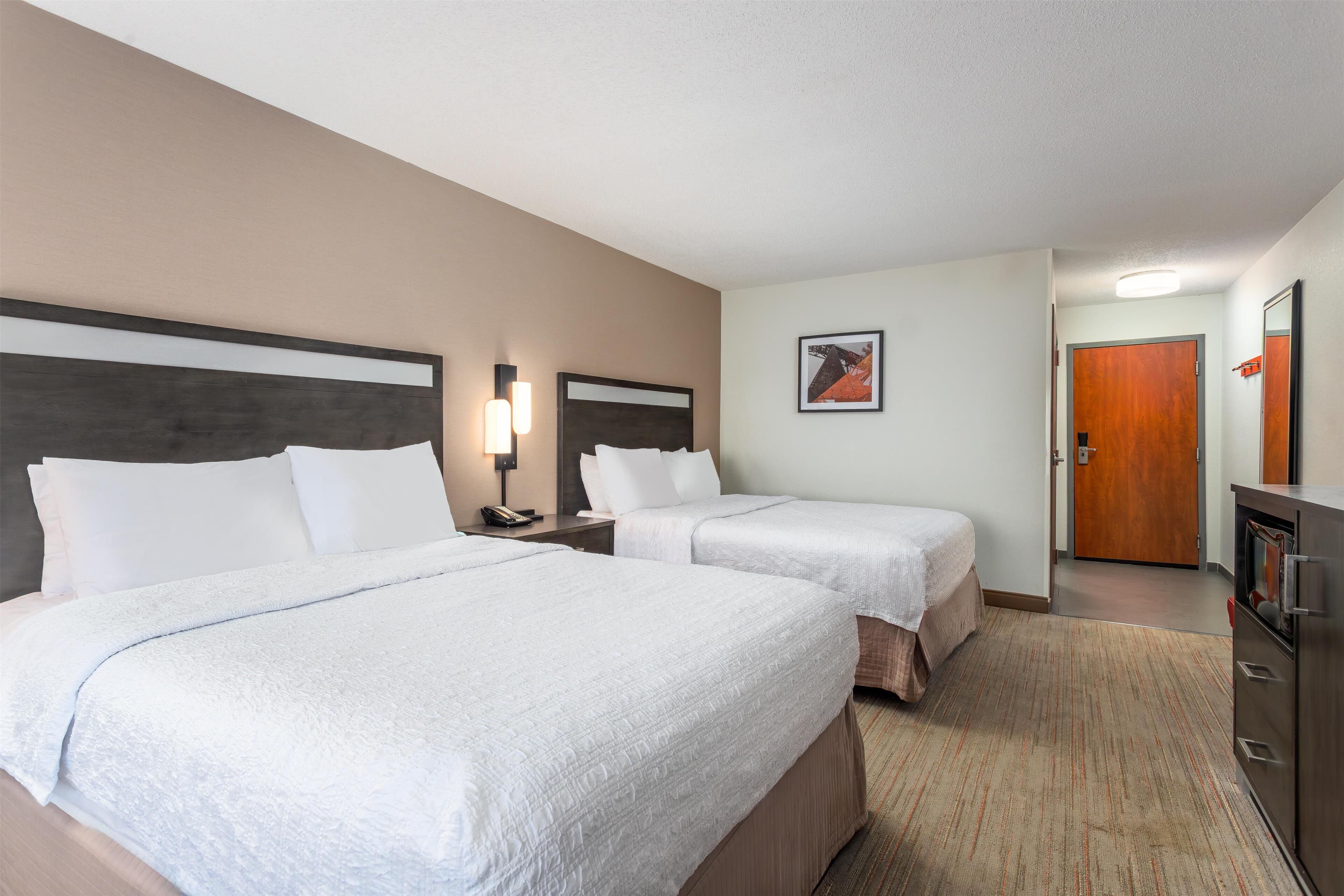 A hotel room with two beds, a wooden headboard, a nightstand with a lamp, a framed picture on the wall, and a wooden door.