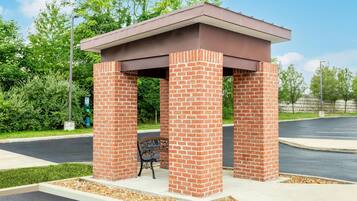 A brick pavilion with a bench, surrounded by greenery and a parking area.