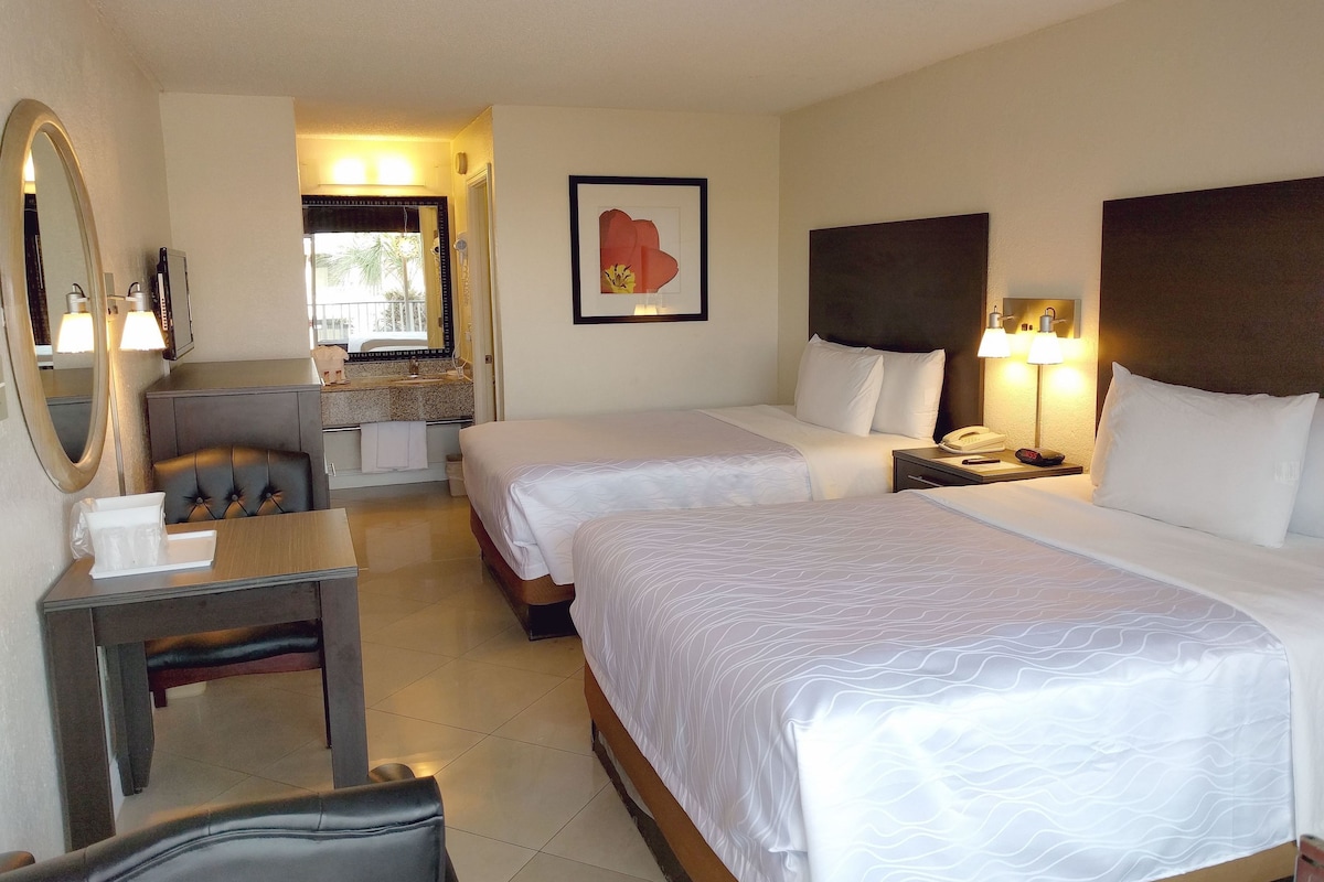 A hotel room with two single beds, a desk with a chair, a vanity with a mirror, and a framed artwork on the wall.
