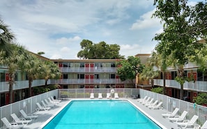 A swimming pool with white lounge chairs, a multi-story building with balconies, and surrounding greenery.