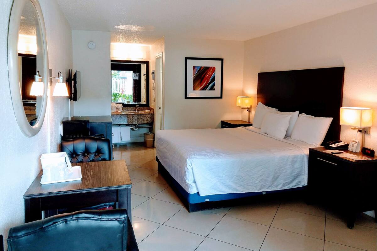 A hotel room with a bed, bedside tables, a desk, a chair, a mirror, a TV, and a bathroom visible through an open door.