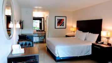 A hotel room with a bed, bedside tables, a desk, a chair, a mirror, a TV, and a bathroom visible through an open door.