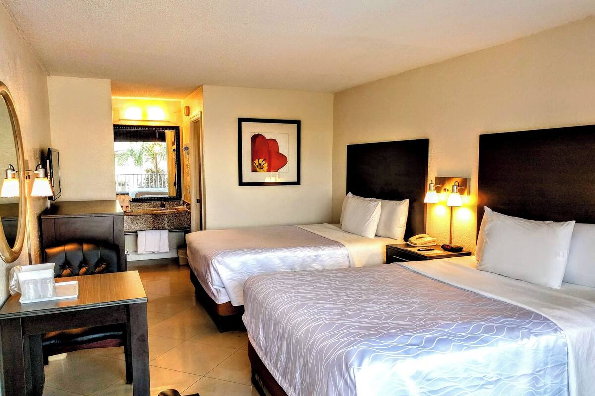 A hotel room with two beds, a desk, a chair, a mirror, a bathroom with a shower, and a framed picture on the wall.