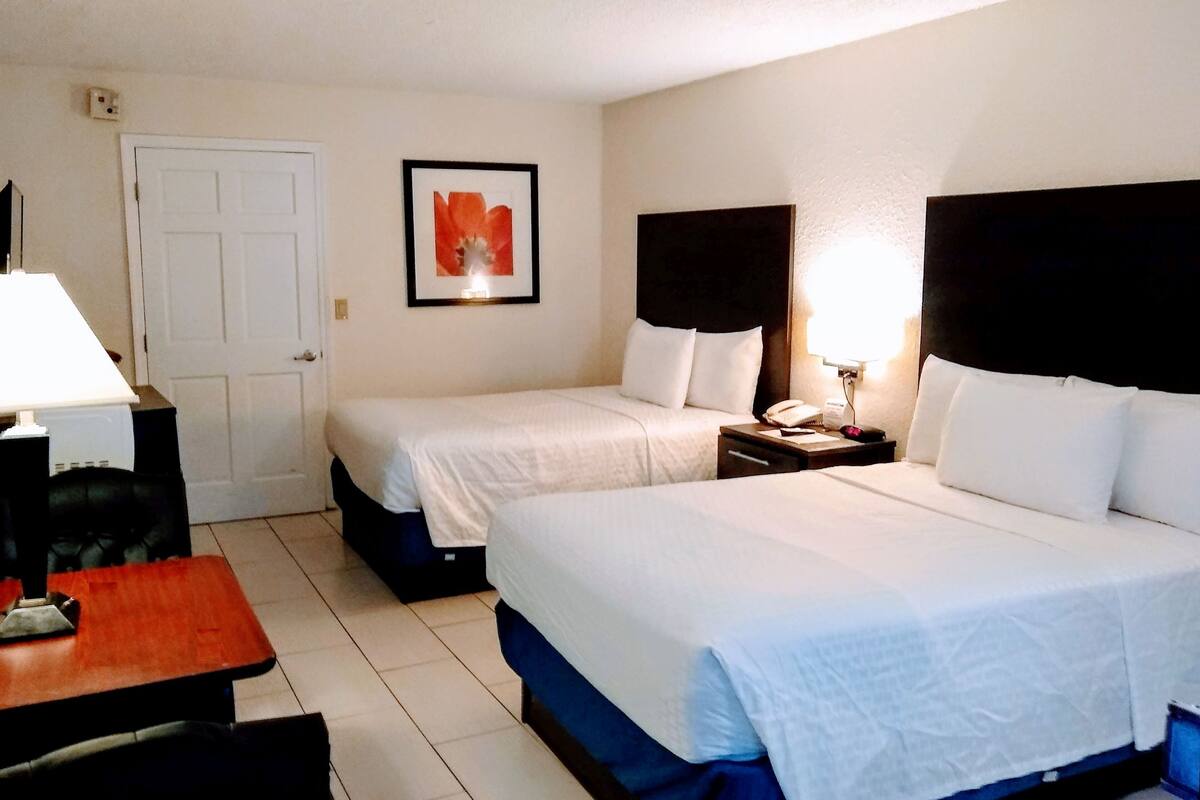 A hotel room with two beds, a desk, a chair, a framed picture on the wall, and a nightstand with a lamp.