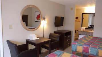 Une chambre d’hôtel équipée d’un bureau, de deux chaises, d’une télévision, d’un miroir, d’un lit recouvert d’une couverture aux couleurs vives, et d’une salle de bain visible à travers une porte ouverte.