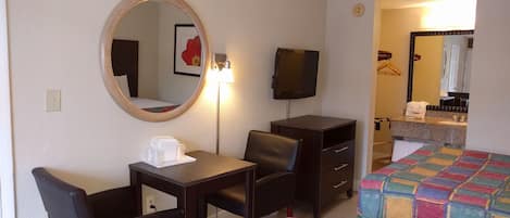 Habitación de hotel con escritorio, dos sillas, televisor, espejo, cama con colcha colorida y baño visible a través de una puerta abierta.