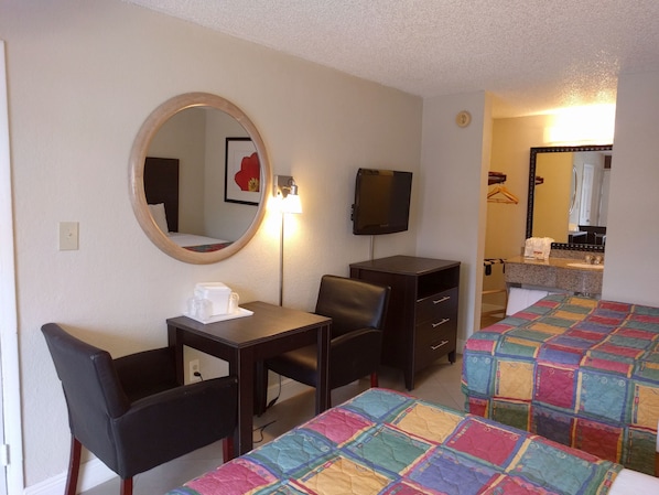 A hotel room with a desk, two chairs, a television, a mirror, a bed with a colorful quilt, and a bathroom visible through an open door.