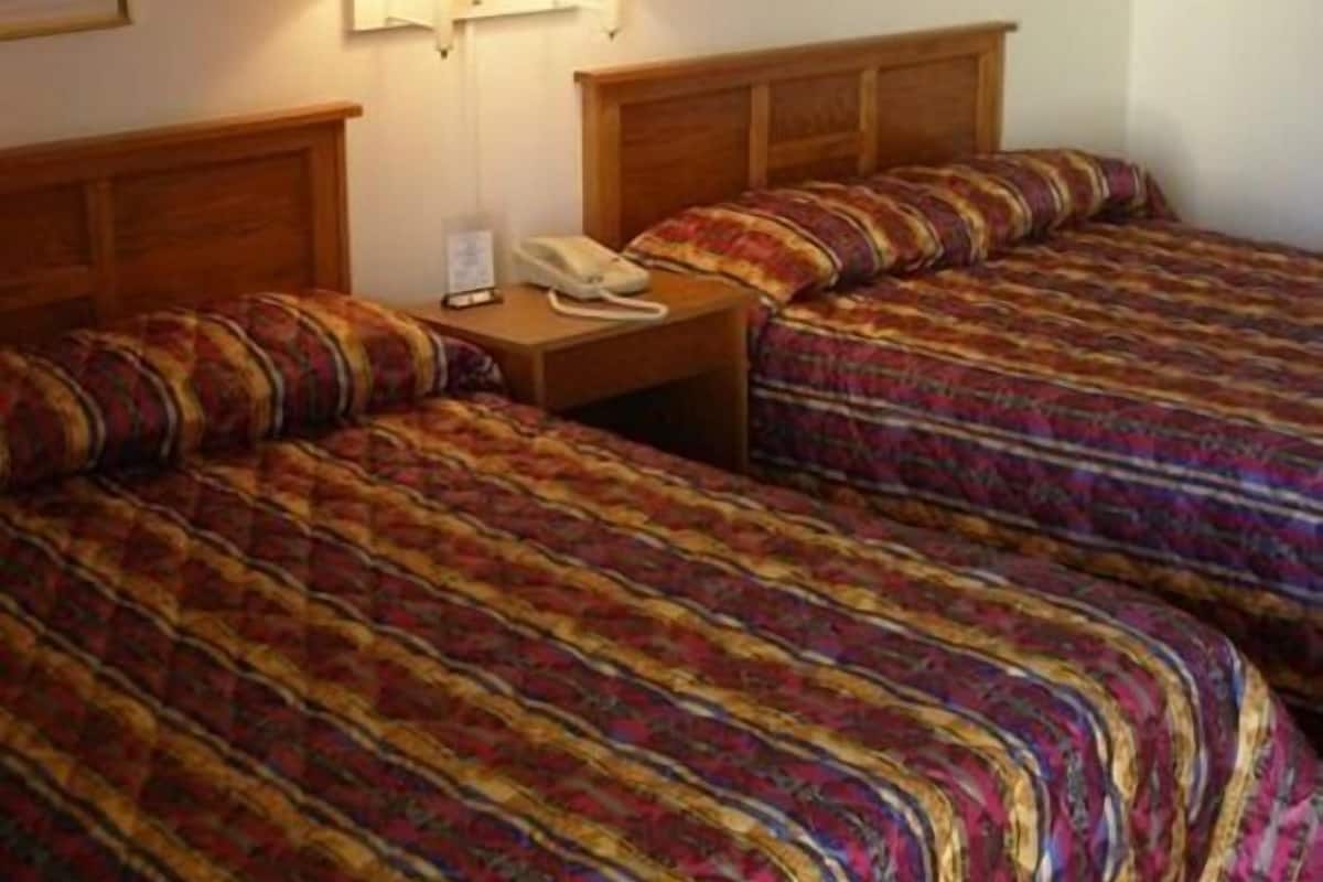 A hotel room with two beds, each with a patterned bedspread. There are two wall-mounted lamps on the nightstand between the beds.