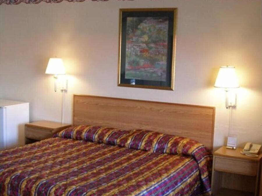 A hotel room with a wooden headboard, a colorful striped bedspread, two bedside lamps, a framed picture on the wall, and a small bedside table with a phone.
