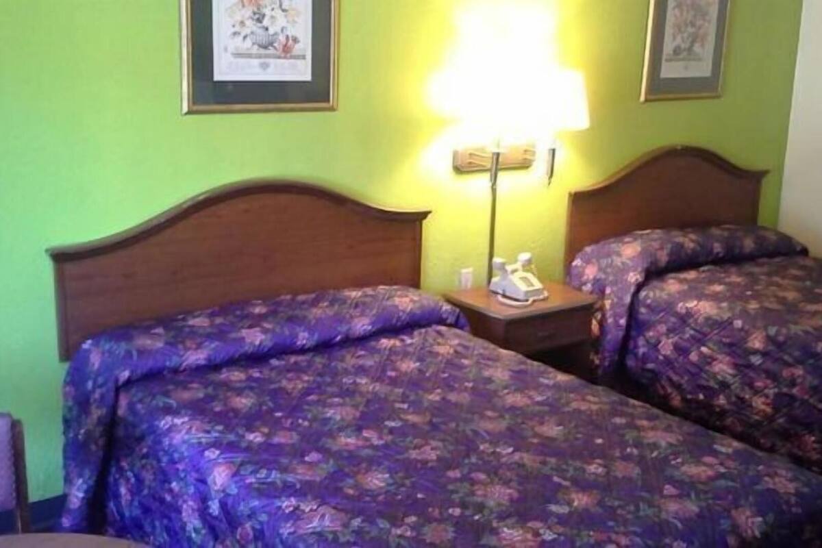 A room with two beds, each with floral bedding, a nightstand with a lamp, and framed pictures on the wall.