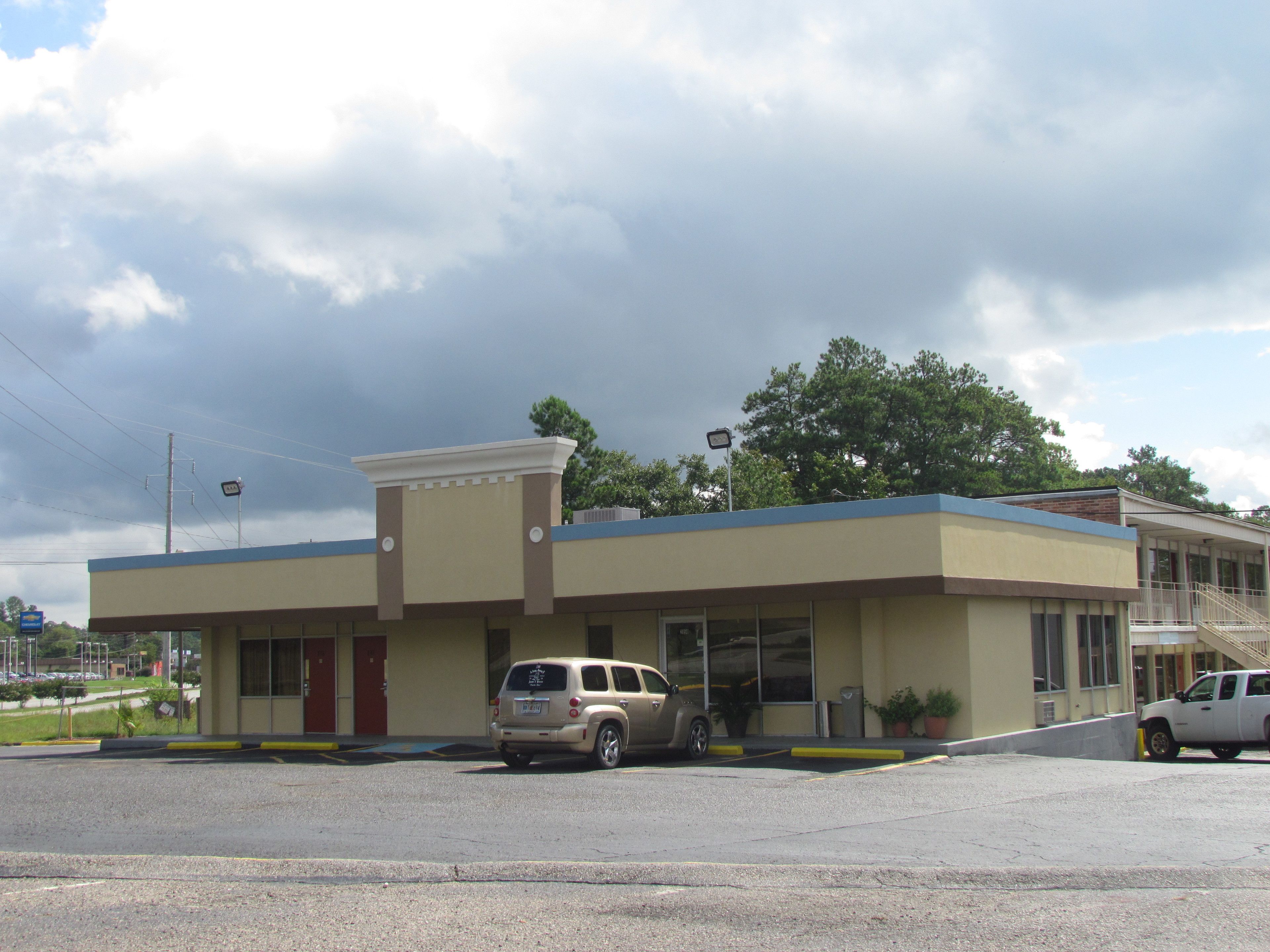 A building with a flat roof, a single-story structure, and a parking lot with two cars.