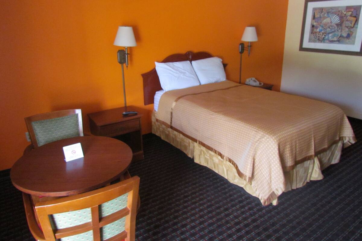 A hotel room with a bed, two bedside lamps, a chair, a small table, and a framed picture on the wall.
