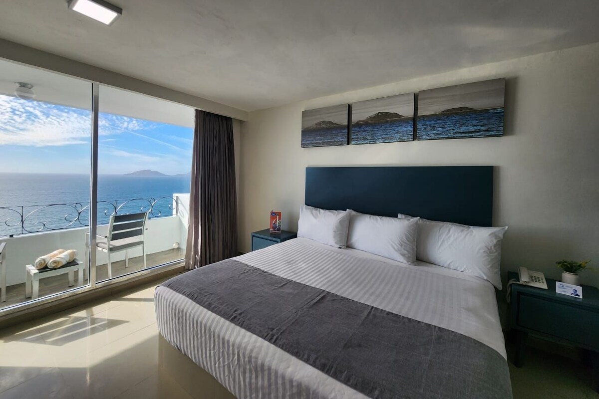 Hotelkamer met een groot bed, nachtkastje, balkon met een stoel en handdoeken, en uitzicht op zee.