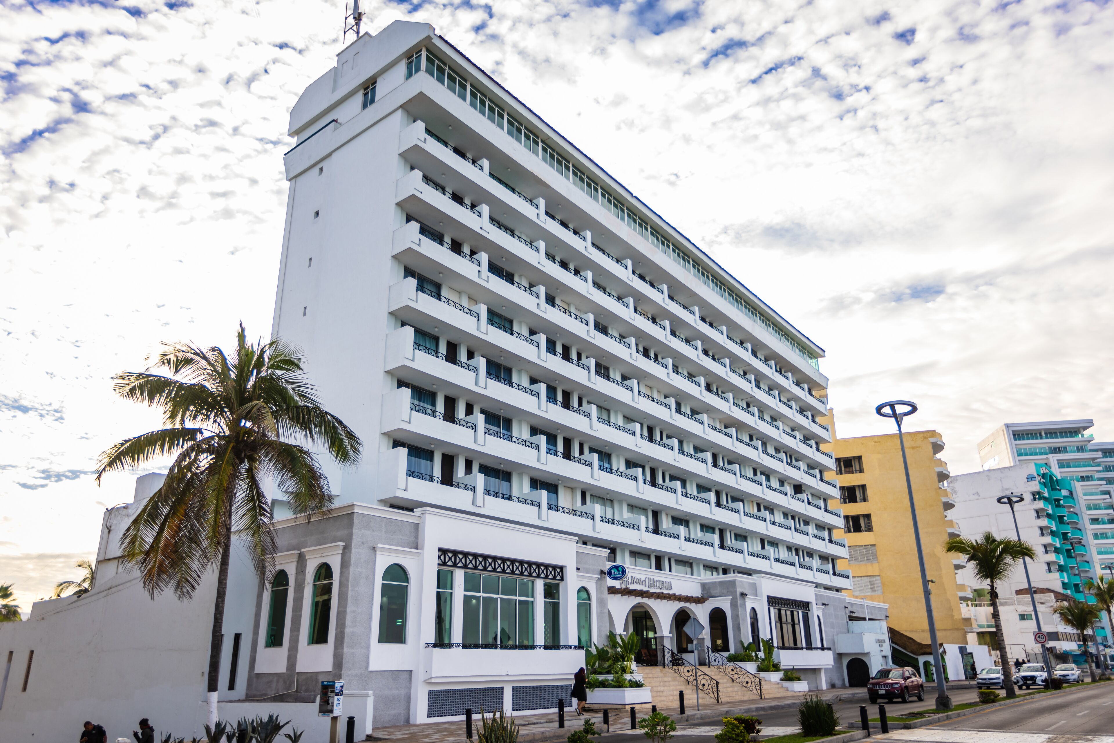 Un hotel de varios pisos con balcones, una entrada central con escaleras y palmeras al frente.
