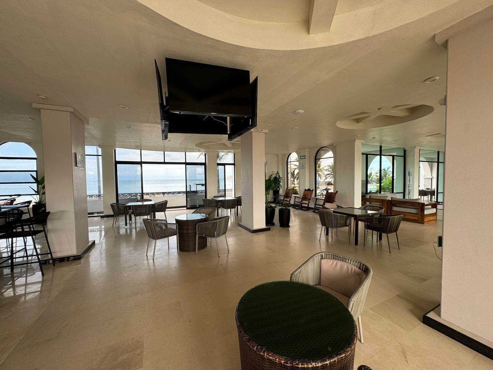 A spacious indoor area with large windows offering views of the ocean, furnished with tables and chairs, and a flat-screen TV mounted on the ceiling.