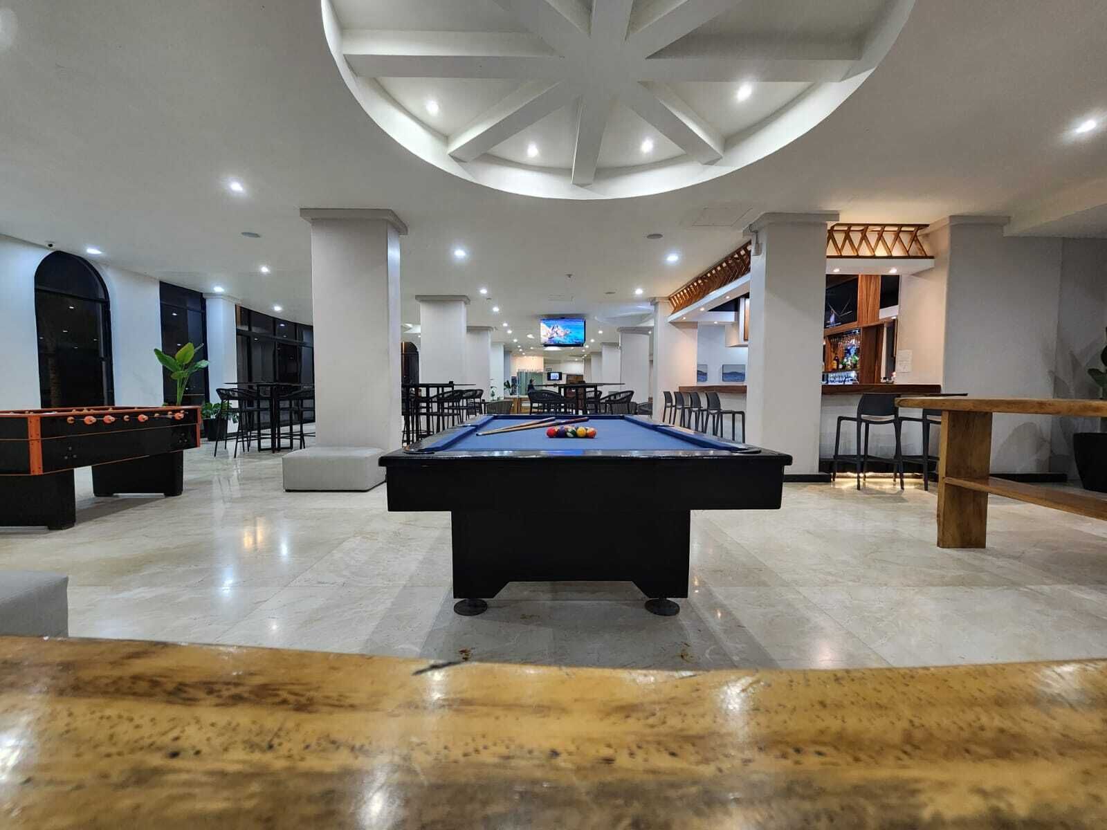 A pool table in a well-lit indoor space with a high ceiling, seating areas, and a bar.