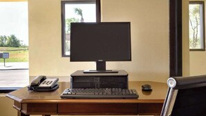 A desk with a computer setup, including a monitor, keyboard, and mouse, in a room with windows and a view of greenery outside. - Travel Inn (Unadilla)
