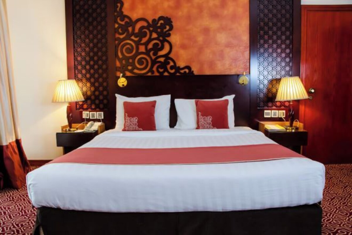 Een hotelkamer met een groot bed, twee nachtkastjes met lampen, een decoratief hoofdbord en een rood tapijt.