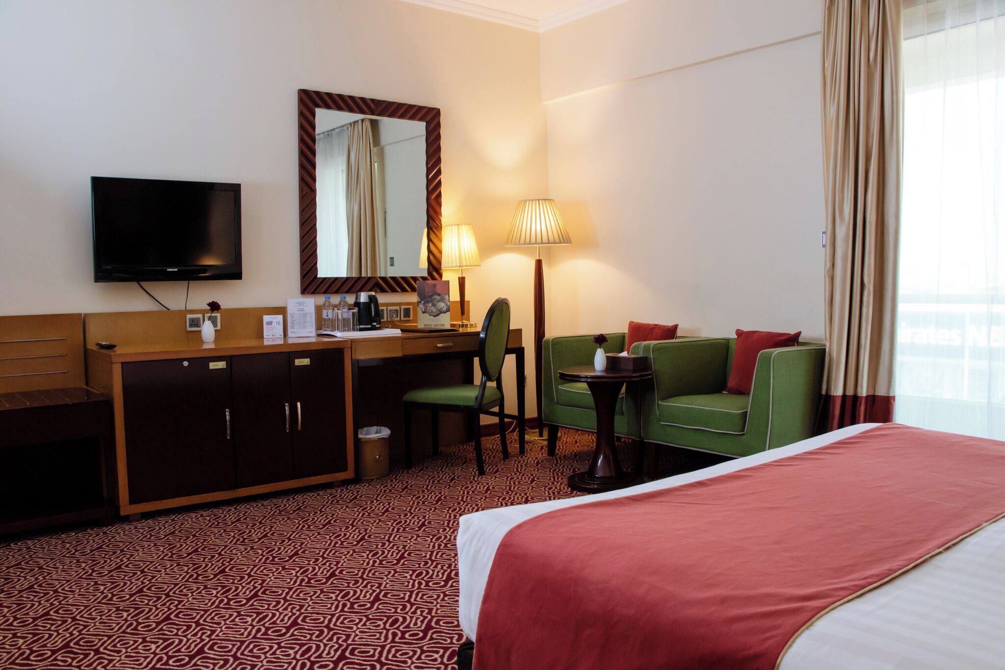 A hotel room with a bed, a desk with a chair, a television, a mirror, a lamp, and a window with curtains.