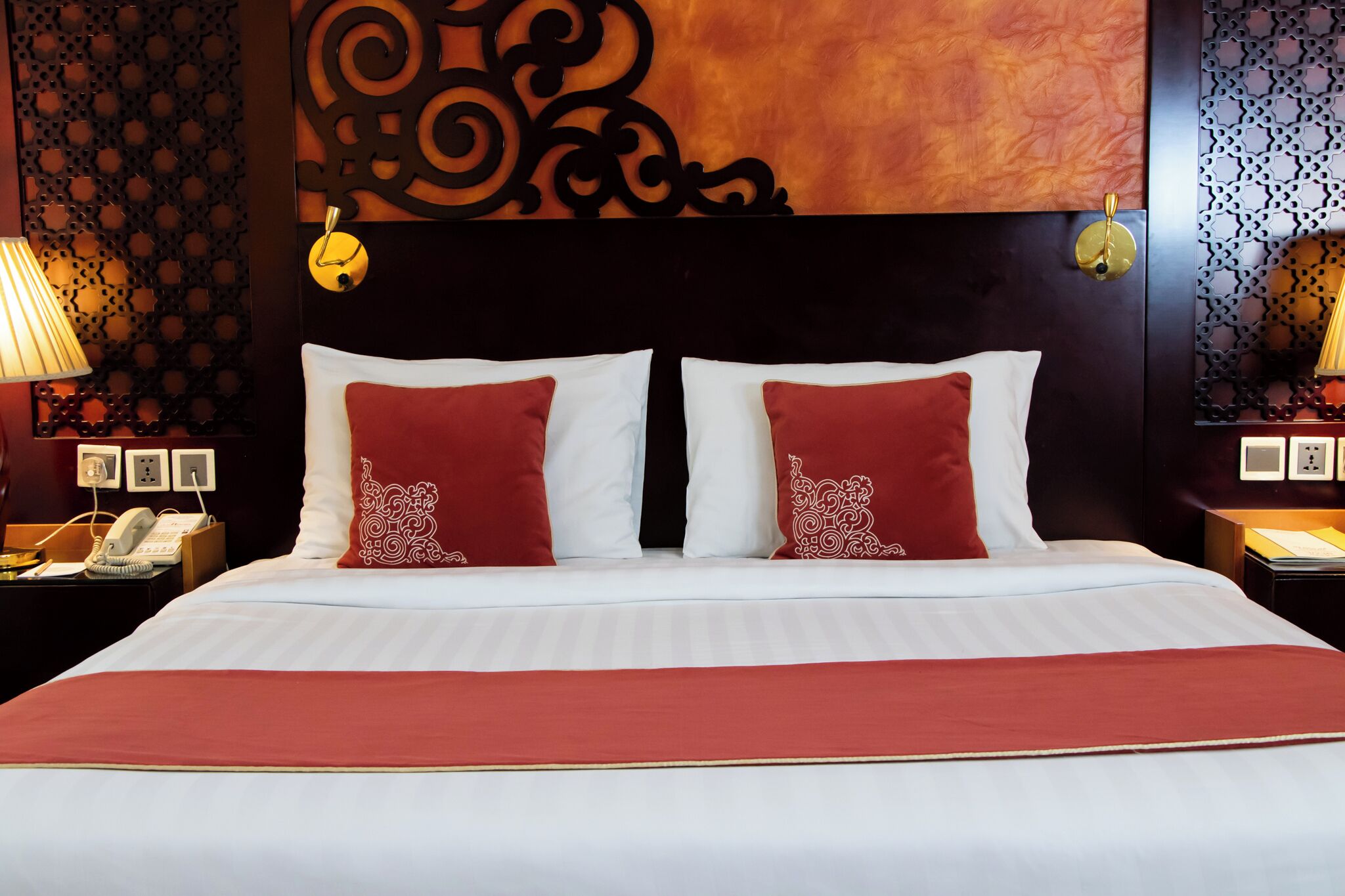 A hotel room with a large bed, two red and white pillows, a red throw blanket, a decorative headboard, and a nightstand with a lamp and phone.