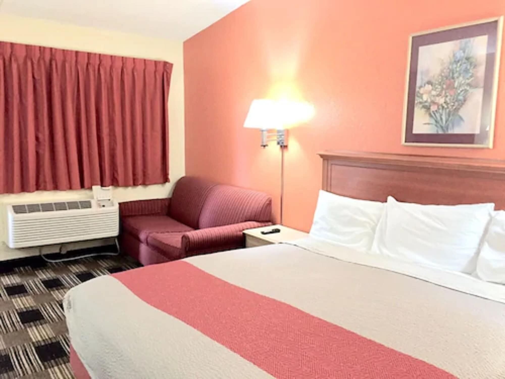A hotel room with a bed, a red chair, a nightstand, a lamp, and a wall-mounted air conditioner.