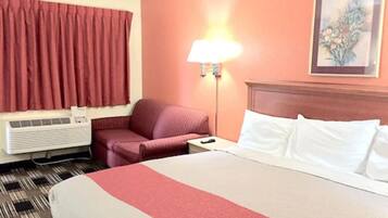 A hotel room with a bed, a red chair, a nightstand, a lamp, and a wall-mounted air conditioner.