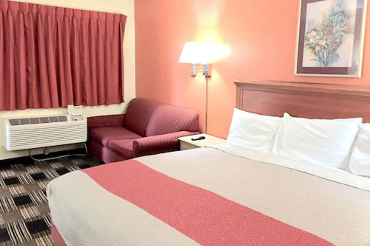 A hotel room with a bed, a chair, a nightstand, a lamp, and an air conditioning unit.