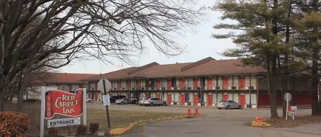 A Red Carpet Inn with a sign indicating the entrance, a parking lot with several cars, and a building with multiple doors and windows.
