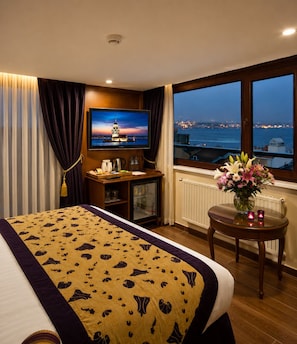A hotel room with a bed, a TV, a desk with a computer, a small table with flowers, and a window with a city view. - GLK PREMIER Acropol Suites & Spa - Special Class  (Istanbul)