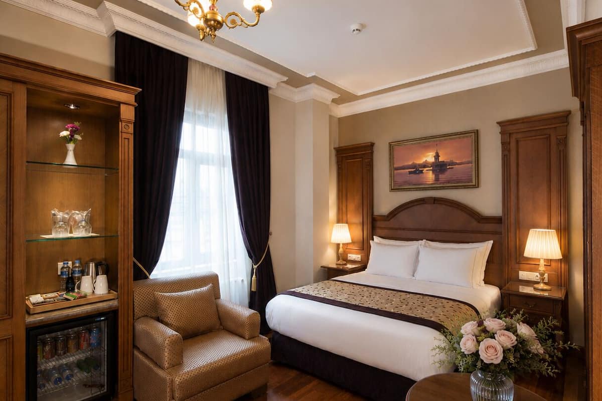 A hotel room with a bed, a chair, a small table, a minibar, and a painting on the wall.