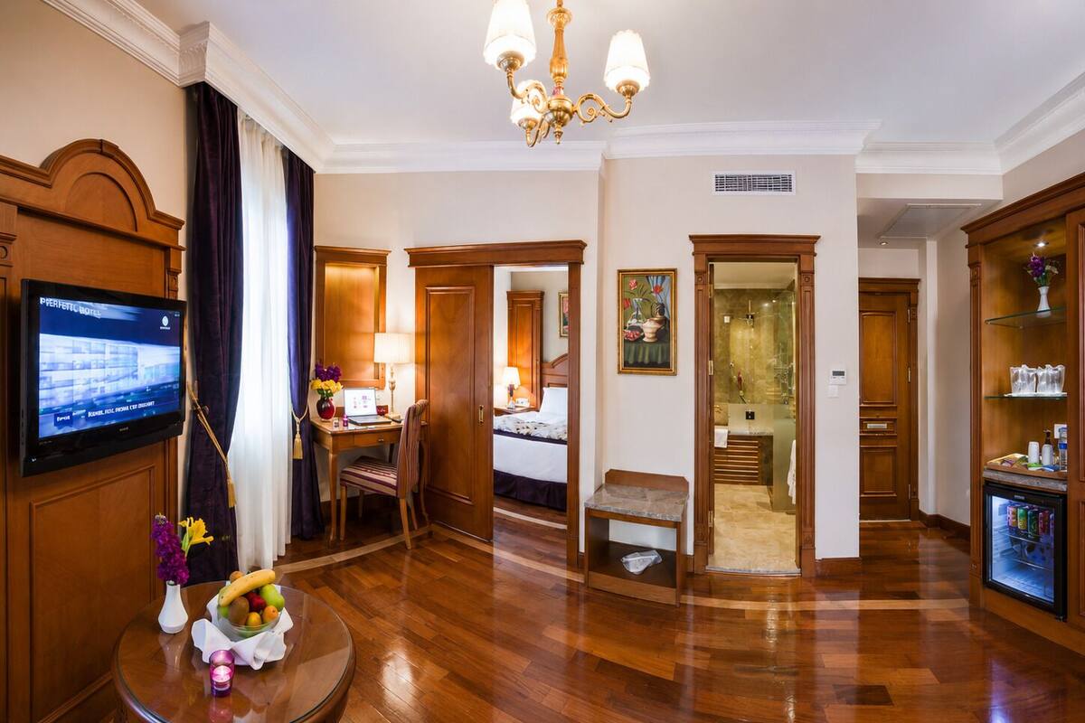 A hotel room with a wooden floor, a desk with a computer, a bed, a bathroom, and a minibar.
