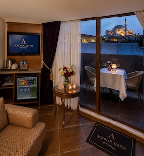A hotel room with a television, minibar, refrigerator, dining table with chairs, and a view of a building with a dome. - GLK PREMIER Acropol Suites & Spa - Special Class  (Istanbul)