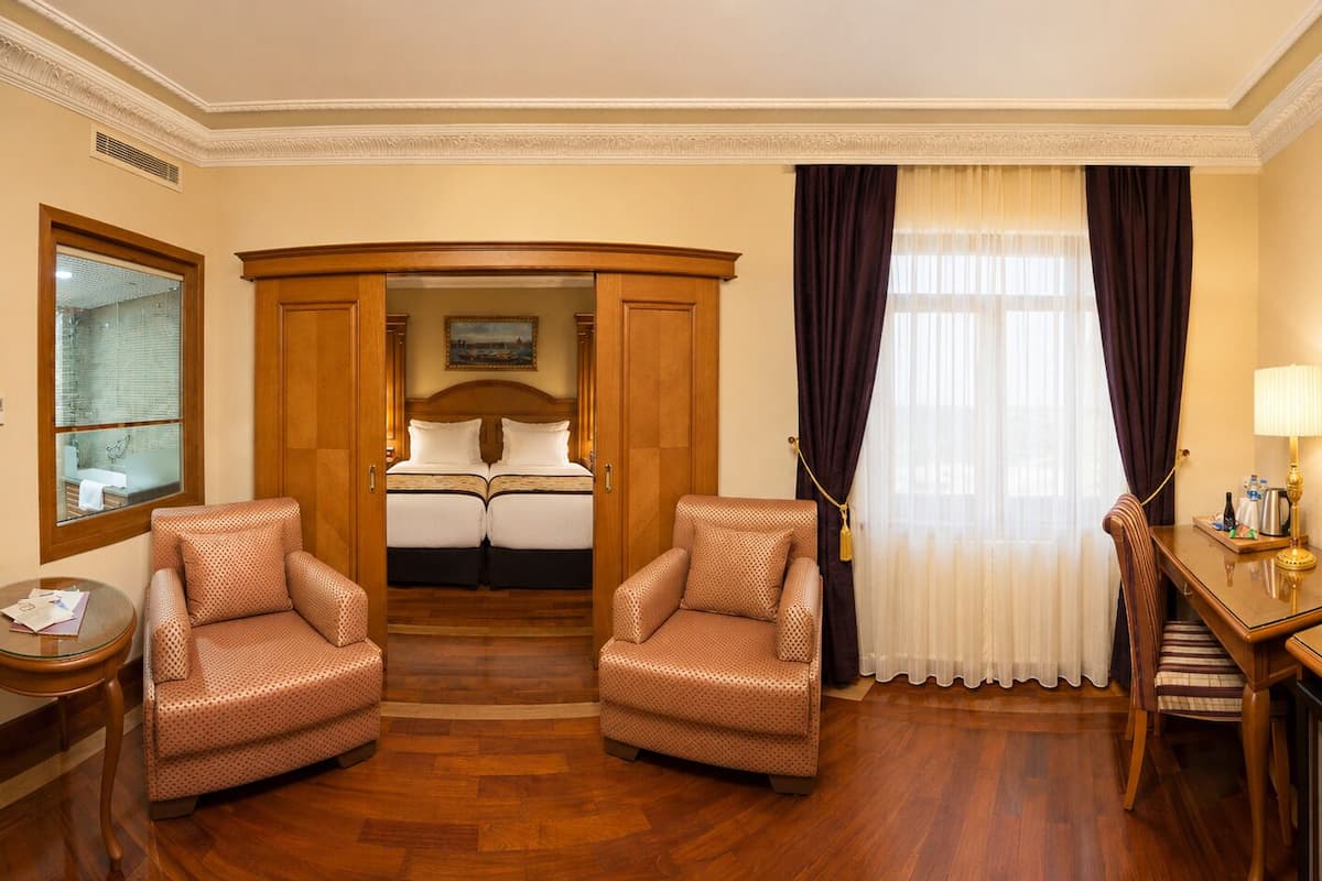 A hotel room with a wooden floor, two armchairs, a bed with white linens, a desk with a lamp, a television, and a window with curtains.