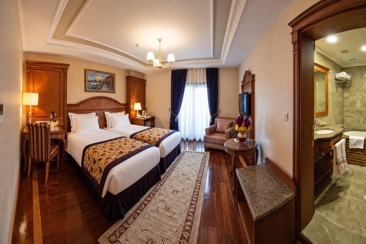 A hotel room with two beds, a sitting area, a desk, a chair, a TV, and a bathroom visible through an open door.