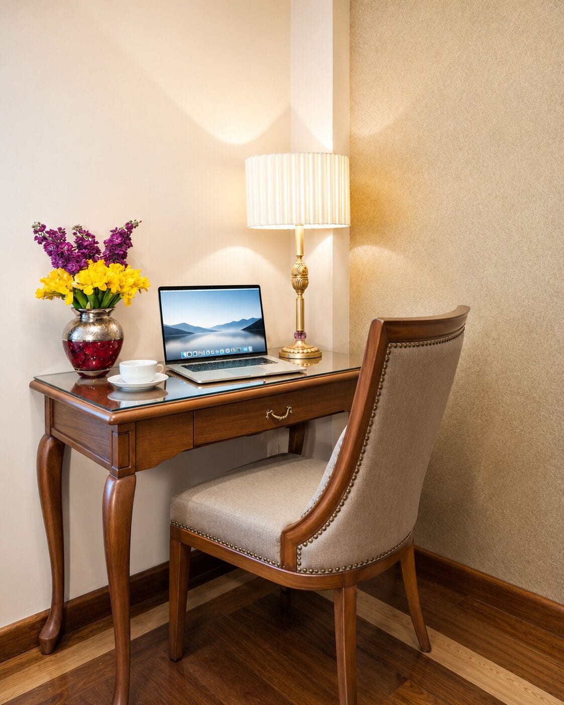 Quarto de hotel com uma escrivaninha de madeira, um laptop, uma luminária, uma cadeira e um vaso com flores.