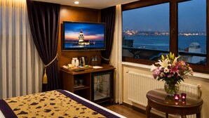 Habitación de hotel con una cama, televisión, escritorio con computadora, una mesita con flores y ventana con vista a la ciudad.
