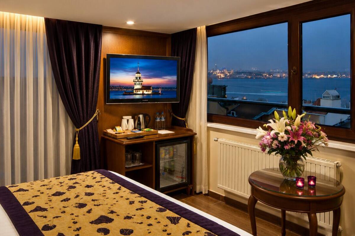 A hotel room with a bed, a TV, a desk with a computer, a small table with flowers, and a window with a city view.
