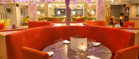 Ein Restaurant mit runden Tischen, orangefarbenen Polstersitzen und einem zentral platzierten Zierbaum.