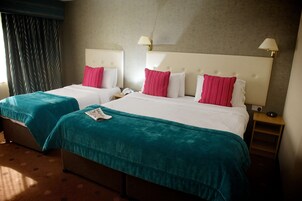 Habitación de hotel con dos camas, ropa de cama azul verdosa y blanca, almohadas rosadas y una mesita de noche de madera con una lámpara.