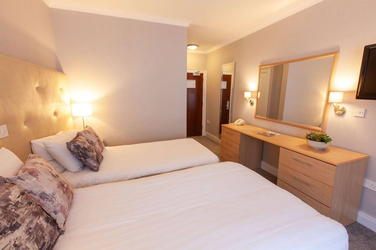 Une chambre d’hôtel avec deux lits simples, une commode en bois, un miroir et des lampes de chevet.