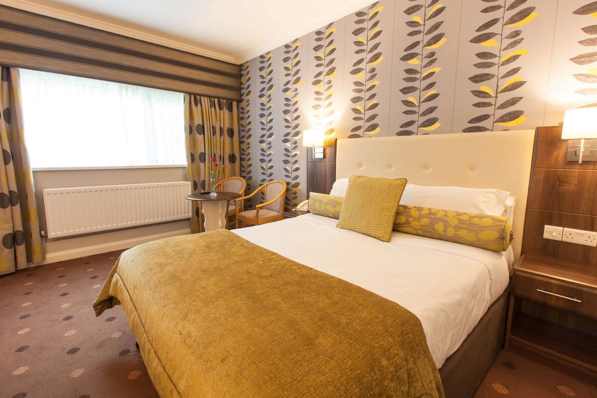 Une chambre d’hôtel avec un grand lit, une table de chevet avec une lampe, une chaise et une fenêtre avec des rideaux.