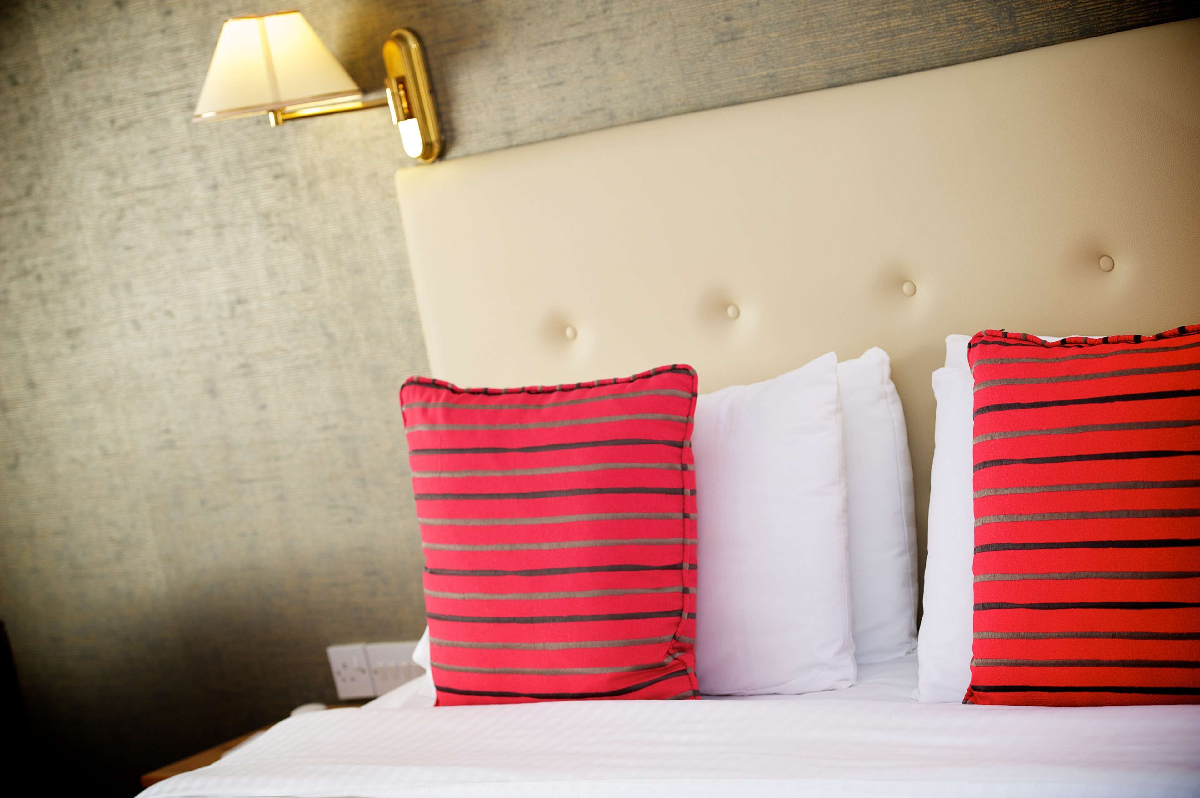 A neatly made bed with white and red striped pillows, a beige headboard, and a wall-mounted lamp.