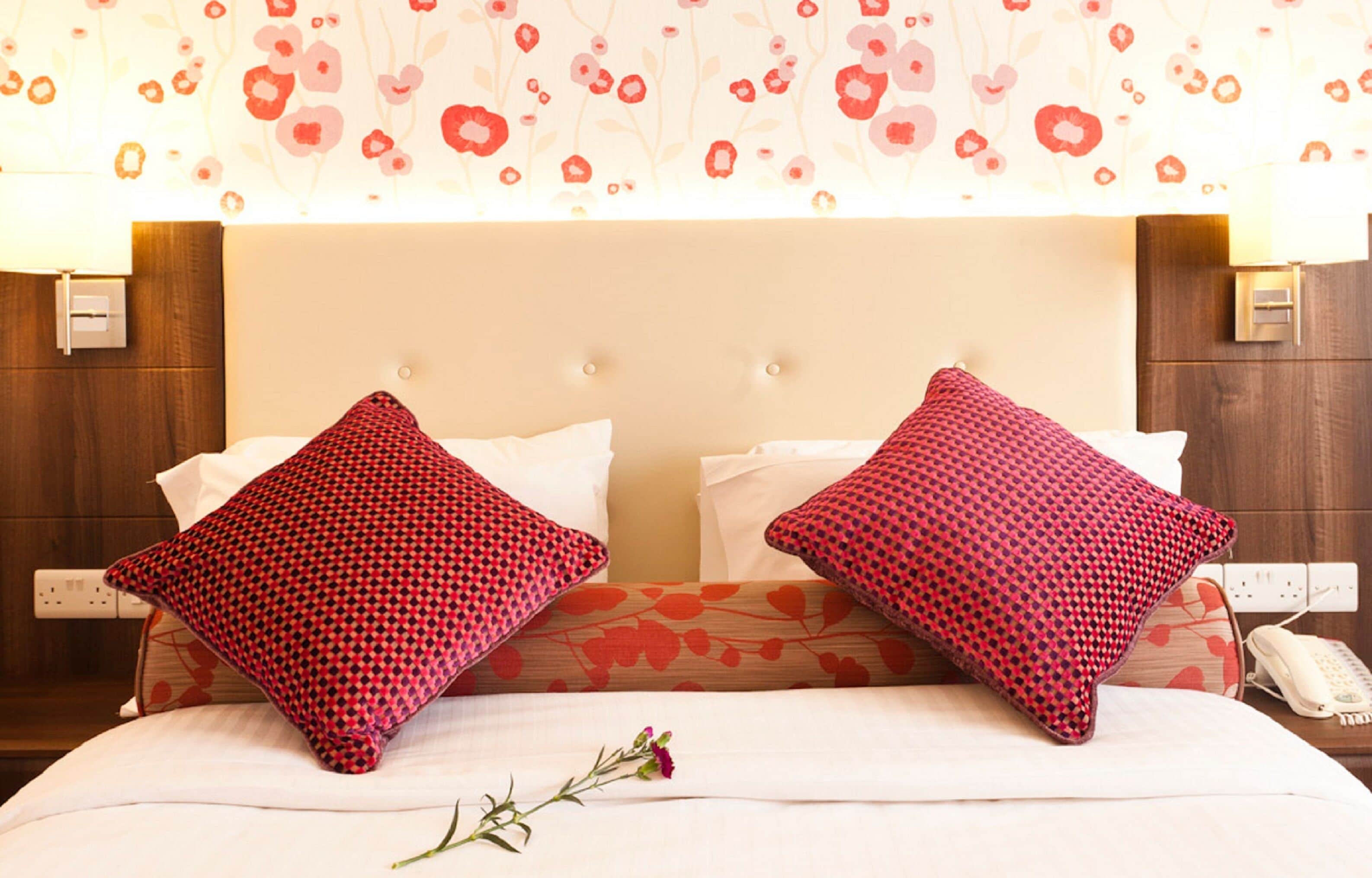 Une chambre d’hôtel avec un lit, deux coussins rouges à motifs, un coussin blanc uni, une table de chevet avec un téléphone et un mur recouvert d’un papier peint à motifs floraux.