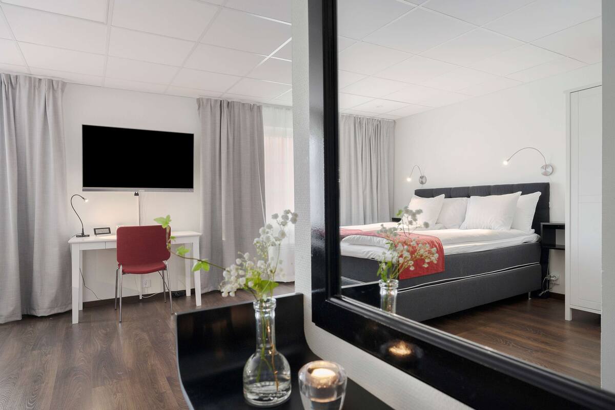 A modern hotel room with a bed, a desk, a chair, a TV, and a mirror reflecting the room.