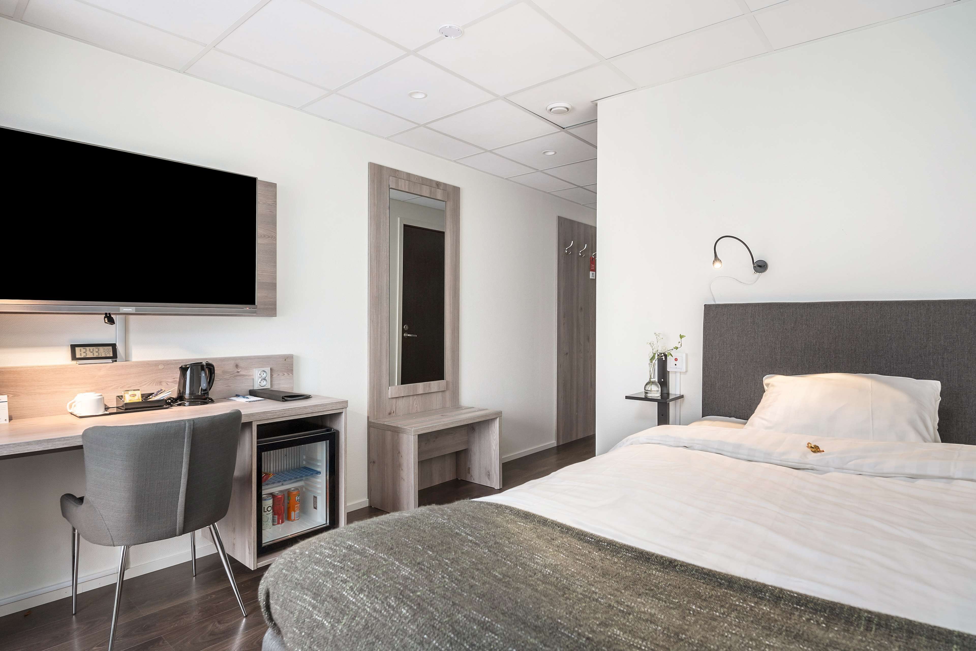 A modern hotel room with a bed, a desk, a chair, a television, and a small refrigerator.