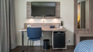 Habitación de hotel con escritorio de madera, una silla azul, un televisor sobre el escritorio, un cubo de basura pequeño y una cama con colcha de rayas azules y blancas.