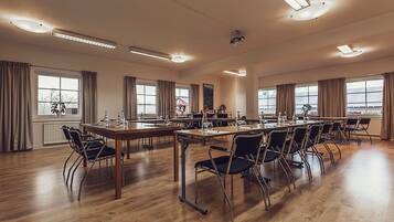A conference room with multiple tables and chairs, large windows with curtains, and ceiling lights.